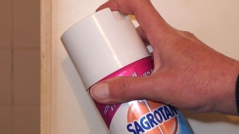 Ein Hand hält eine Sagrotan-Sprühflasche, um Klebereste auf Kunststoff zu entfernen, während sich eine Wand im Hintergrund befindet.