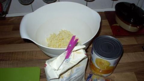 In einer weißen Schüssel wird Käse gerieben. Daneben stehen Mehl, eine Dose mit Pfirsichen und Vanillepudding auf der Küchentheke.