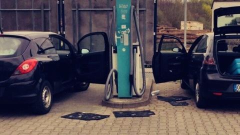 Zwei Autos stehen an einer Tankstelle, während Teppichreste als Bodenmatten im Außenbereich liegen.