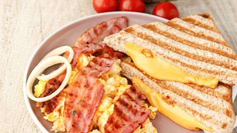 Gegrilltes Käse-Sandwich liegt auf einem Teller neben Rührei und gebratenem Speck, umgeben von Kirschtomaten und Zwiebelringen.