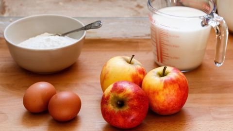 Auf einem Holzbrett liegen &Auml;pfel, Eier und eine Sch&uuml;ssel mit Mehl, w&auml;hrend ein Messbecher Milch bereitsteht.