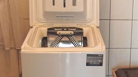 Eine Toplader-Waschmaschine mit offenem Deckel zeigt ein beschädigtes Metallfach, in dem ein Plastiklöffel liegen könnte.