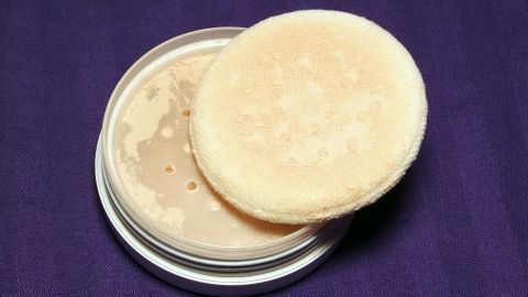 Ein Puder und ein Applikator liegen offen in einer runden Dose auf einem lila Hintergrund, ideal für S.0.S-Make-up.