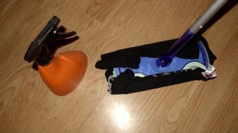 Ein Bodenwischer liegt auf einem Holzfußboden, daneben steht eine orangefarbene Sprühflasche für die Reinigung.