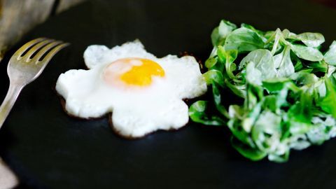 Ein gedünsteter Feldsalat liegt neben einem Spiegelei, das wie eine Blume aussieht, auf einem schwarzen Teller.