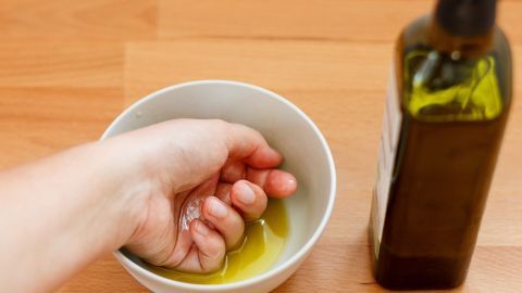 Eine Hand mit rissigen Fingern taucht in eine Schüssel mit Öl, während eine Flasche Olivenöl daneben steht.