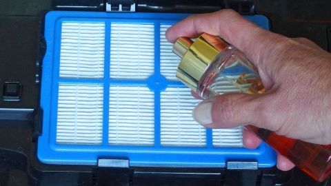 Tipp gegen schlechte Luft aus dem Staubsauger: Staubsaugerfilter mit Parfum besprühen. Ein Hand hält eine Sprühflasche über einen blauen Staubsaugerfilter, um ihn zu reinigen und die schlechte Staubsaugerluft zu verbessern.