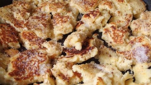 In einer Pfanne liegt ein gebräuntes Quark-Schmarren, das zart zerfällt, umringt von feinem Zucker und Apfelstücken.
