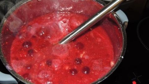 Ein Topf kocht Himbeer-Kirsch-Marzipan-Konfitüre mit dampfender roter Masse und ein paar Kirschen auf dem Herd.