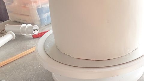 Ein runder, weißer Fondantkuchen steht auf einem Drehständer, umgeben von Werkzeugen und Material zum Dekorieren.