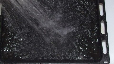 Ein Backblech wird mit Scheuermilch gereinigt, während Wasser darüber spritzt, um Schmutz zu entfernen.