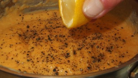 Eine Hand drückt eine Zitronenhälfte über einer cremigen Karotten-Kartoffelsuppe, die mit schwarzem Pfeffer gewürzt ist.