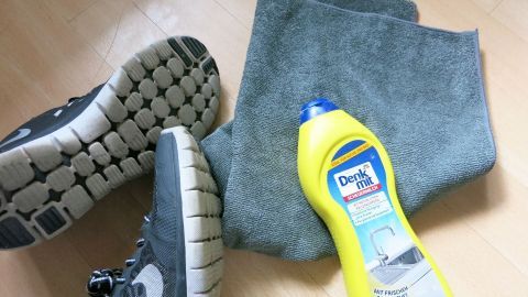 Schuh neben einem grauen Tuch und einer Flasche "Denk mit Scheuermilch" auf Holzboden; geeignet zum Reinigen und Pflegen.