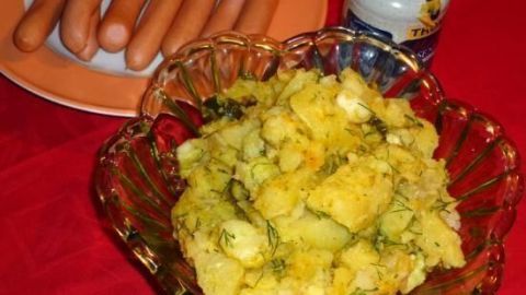 Ein bunter Kartoffelsalat in Dill-Marinade liegt in einer Schüssel, während daneben Würstchen auf einem Teller warten.