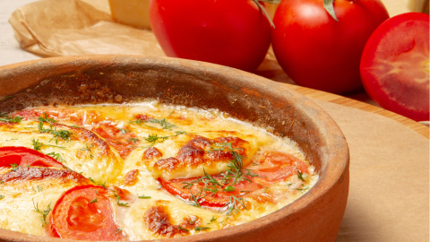 Ein köstlicher Grünkernauflauf mit frischen Tomatenscheiben und Kräutern in einem rustikalen Tonbehälter, umgeben von reifen Tomaten.