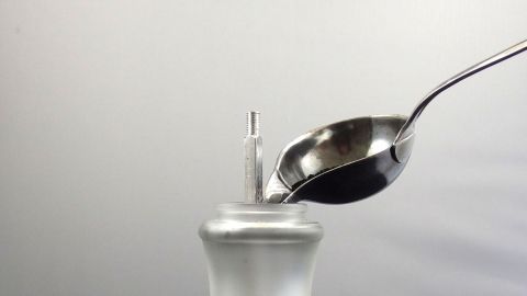 Eine handliche Pfeffermühle wird mit einem Löffel gefüllt, während sie vor einem neutralen Hintergrund steht.