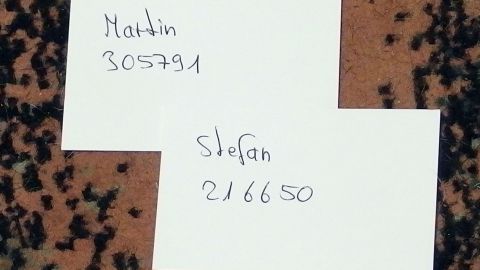 Kleine Zettel liegen auf einem Teppich, auf denen Namen und Zahlen handgeschrieben sind: „Martin 305791“ und „Stefan 216650“.