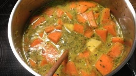 Ein Topf mit Kürbissuppe kocht auf einem Herd. Große orangefarbene Kürbisstücke schwimmen in einer grünen Brühe.