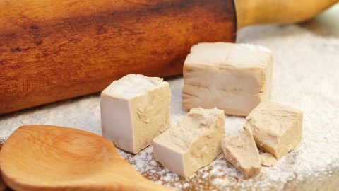 Würfel aus Backhefe liegen auf einer Mehl-unterlegten Arbeitsfläche, neben einem Holzrollingpin und einem Löffel.