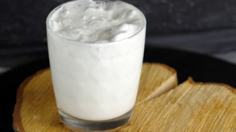 Ein Glas mit aufgeschäumter Milch steht auf einem Holzbrett, umgeben von einer dunklen, neutralen Umgebung.