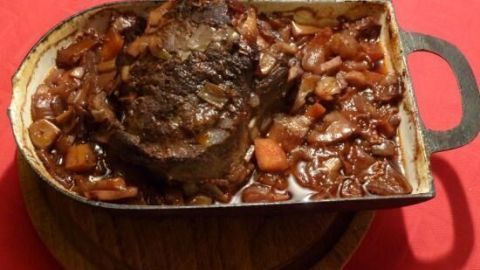 Ein geschmortes Ochsenbäckchen liegt in einer metallenen Auflaufform, umgeben von schmackhaftem Gemüse und Sauce auf einem Holzbrett.