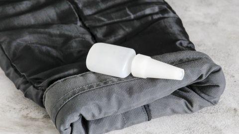 Mit etwas Textilkleber kannst du zu lange Jackenärmel im Handumdrehen kürzen - ganz ohne nähen! Ein grauer Jackenärmel liegt flach auf einer Oberfläche, während eine Flasche mit Flüssigkeit daneben positioniert ist.