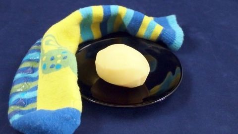 Eine heiße Kartoffel in einem Socken kann ein einfaches und effektives Mittel gegen Halsschmerzen sein. Der Artikel erläutert, wie du diese Hausmittel nutzen kannst, um schnell Linderung zu finden. Eine heiße Kartoffel liegt auf einem schwarzen Teller, während ein bunter, gestrickter Socken daneben liegt.