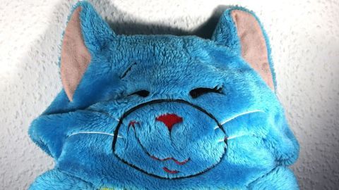 Ein blaues Stofftier mit einem lächelnden Gesicht liegt auf einem hellen, weichen Untergrund und wartet auf eine Umarmung.