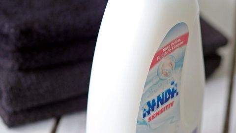 Eine Flasche Weichspüler steht neben einem Stapel dunkler Handtücher auf einem Holzuntergrund.