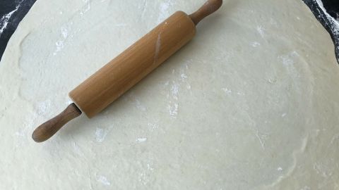 Ein glatter Nudelteig liegt ausgerollt auf einer Arbeitsfläche, während ein Holzrollingpin darauf liegt.