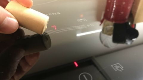 Schwer mit den Fingern bedienbare Glaskeramikfelder lassen sich mit einem Flaschenkorken bedienen. Eine Hand hält einen kleinen Gegenstand über der Touchzone eines Glaskeramikherds, auf dem eine rote Kontrollleuchte leuchtet.