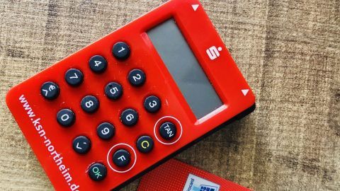 Ein rotes TAN-Gerät liegt auf einer Holzoberfläche neben einer Karte, bereit zur Nutzung für das Sicherheitsverfahren Smart TAN Optic.