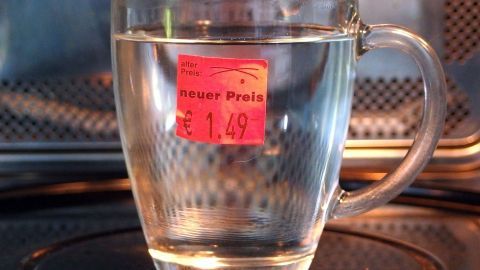 Ein Glas Wasser steht in einer Mikrowelle, auf der ein hartnäckiges Etikett geklebt ist, das den Preis zeigt: 1,49.