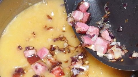 Eine warme Kartoffelsuppe wird aus einem Topf geschöpft, wobei sie mit Speckwürfeln und Zwiebeln angereichert wird.