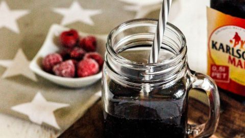 Ein Glas mit dunklem Getränk und einem Strohhalm steht auf einem Holzteller, umgeben von Himbeeren und einer Flasche.