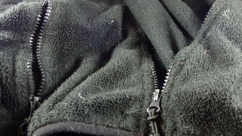 Eine schwarze, flauschige Jacke mit zwei offenen Reißverschlüssen liegt zusammengefaltet auf einem Hintergrund.