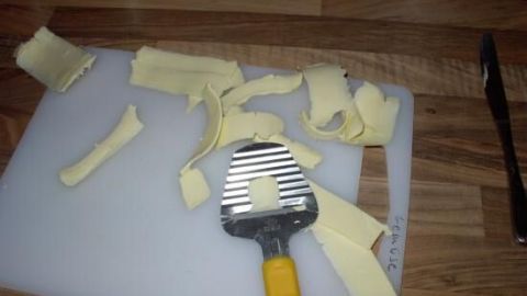 Auf einem hellen Schneidebrett liegt gefrorene Butter, die mit einem Spatel in schmale Streifen geschnitten wird.