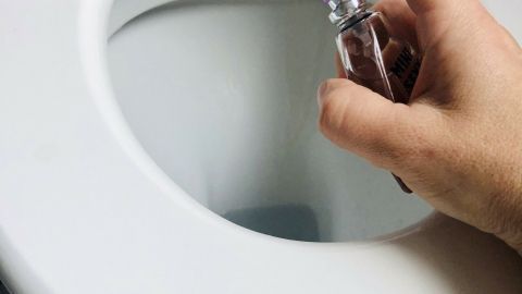 Eine Hand sprüht Eau de Cologne in die Toilette, um einen Duft im Bad zu erzeugen, umgeben von weißen Fliesen.