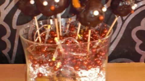 Rentier Cake-Pops auf Stäbchen mit Brezelgeweihen stecken in einem Glas, gefüllt mit roten Perlen, auf Holztisch.