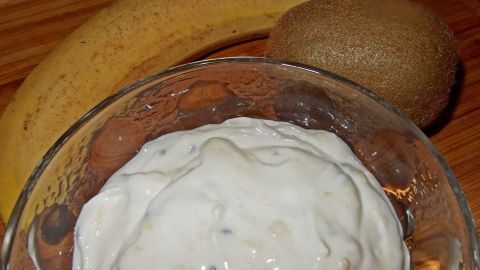 Eine Schüssel mit cremigem Fruchtjoghurt steht auf einem Holztisch, umgeben von einer Banane und einer Kiwi.