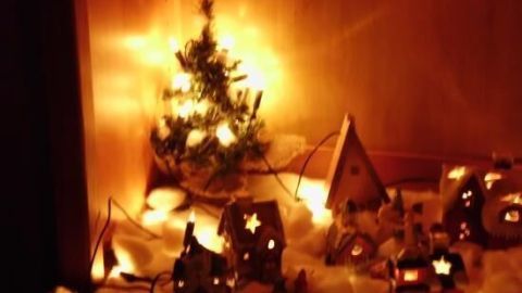 Miniaturhäuser leuchten in einem Weihnachtsdorf, umgeben von Kunstschnee. Ein beleuchteter Baum steht in der Ecke.