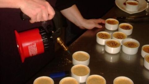 Eine Person karamellisiert Creme Brulee mit einem roten Flambierbrenner in einer Küche, umgeben von diversen Ramekins auf dem Tisch.