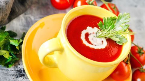 Eine gelbe Tasse mit schneller Tomatensuppe, garniert mit Sahne und frischen Kräutern, steht auf einem Teller, umgeben von Tomaten.