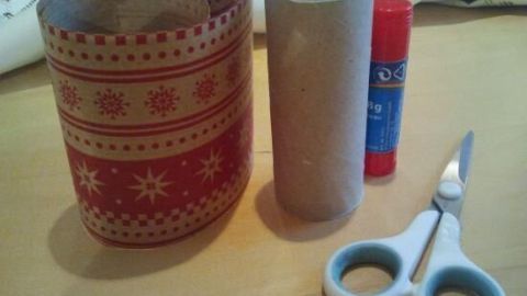 Eine schöne, weihnachtlich dekorierte Geschenkverpackung steht neben einem leeren Papprollenkörper, Kleber und Schere auf einem Tisch.