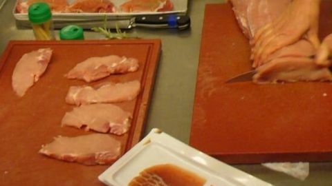 Ein Stück Schweinefleisch wird auf einem großen Schneidebrett in dünne Scheiben geschnitten, um Saltimbocca zuzubereiten.