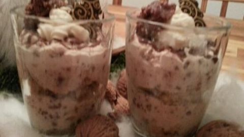 Zwei Gläser gefüllt mit Schichtdessert, dekoriert mit Sahne und Schokolade, stehen auf einem Tisch, umgeben von Walnüssen.