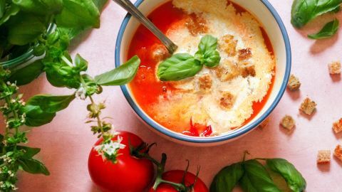 Eine Schüssel frische Tomatensuppe mit Basilikum und Croutons wird von einem Löffel umrührt, umgeben von Tomaten und Kräutern.