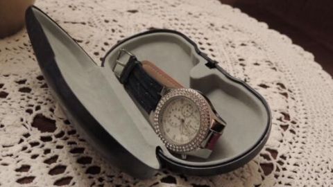 Ein offenes Brillenetui mit einer Armbanduhr darin liegt auf einem gehäkelten Tischdeckchen.