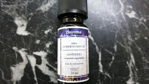 Lavendelöl kann bei Ohrenschmerzen helfen. Ein paar Tropfen, leicht angewärmt, um das Ohr aufgetragen, wirken lindernd und beruhigend. Das Bild zeigt eine Flasche Lavendelöl, das in diesem Hausmittel verwendet wird. Eine Flasche Lavendelöl mit schwarzem Deckel steht auf einem marmorierten Untergrund. Text: "pajoma, 100% ÄTHERISCHES ÖL, LAVENDEL, Lavandula angustifolia, Zart & romantisch, INHALT 10 ml".