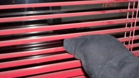 Ein Hand mit einem schwarzen Tuch reinigt die roten Jalousien, während sie in einem Raum mit gedämpftem Licht hängen.
